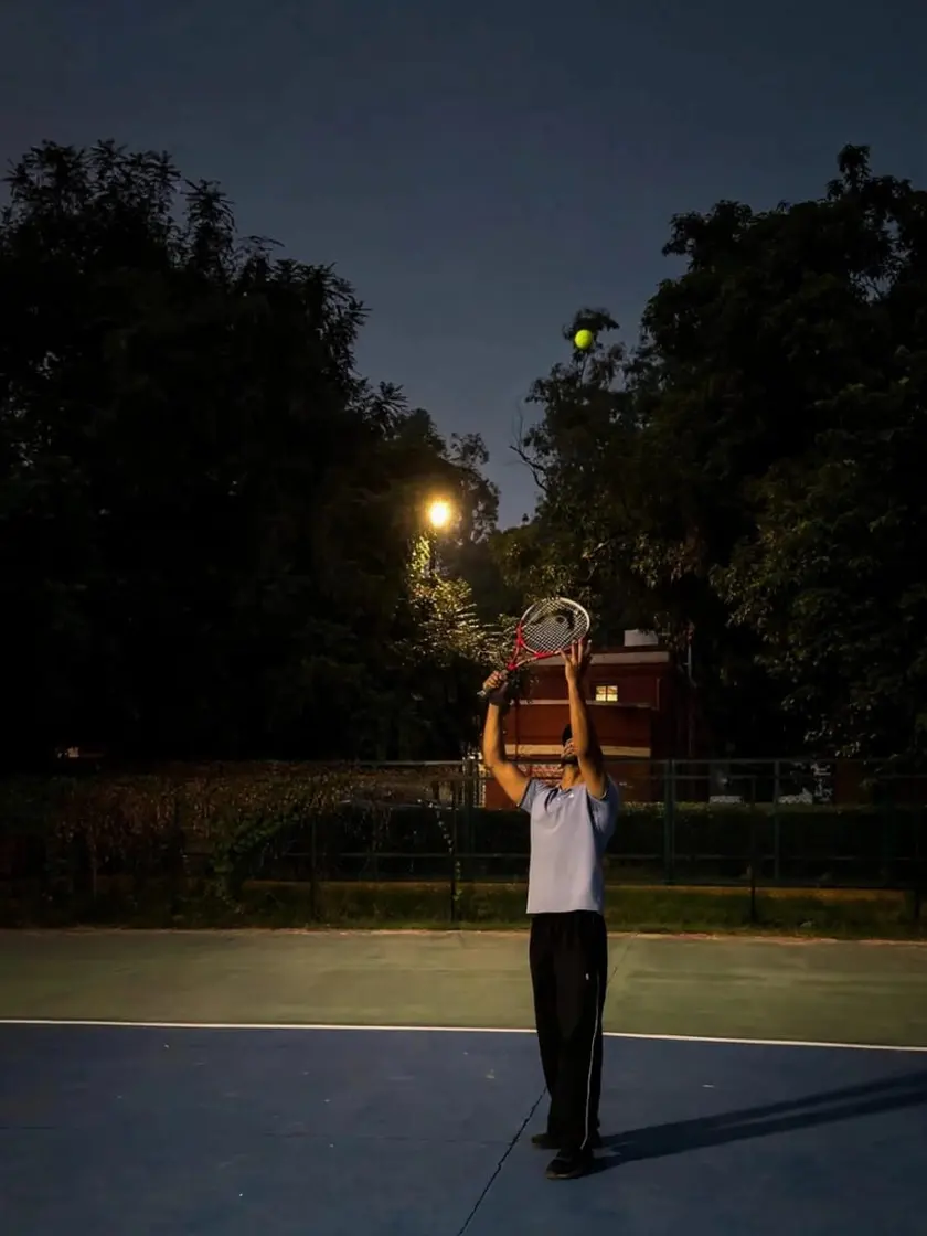 Yuvi mid serve on a floodlit tennis court at dusk, racket raised and the ball in the air.